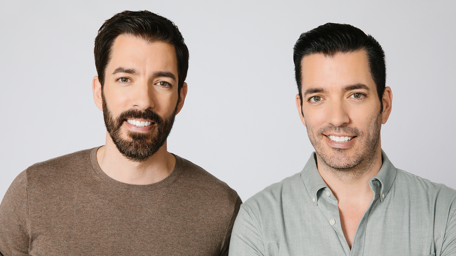 Two men smiling and posing together against a light gray studio background. The man on the left wears a brown crewneck sweater, and the man on the right wears a light sage green button-down shirt.