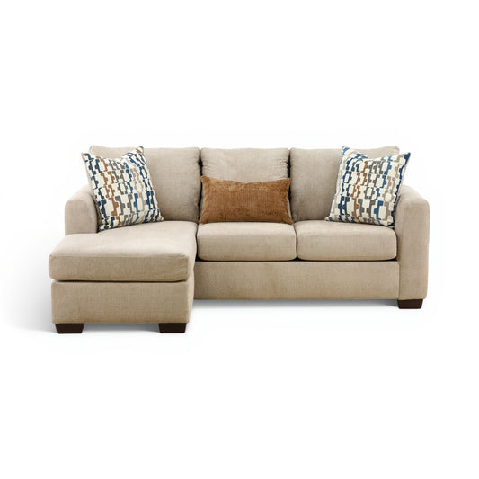 Parchment sofa chaise with chaise ottoman and two patterned throw pillows over beige cushions.