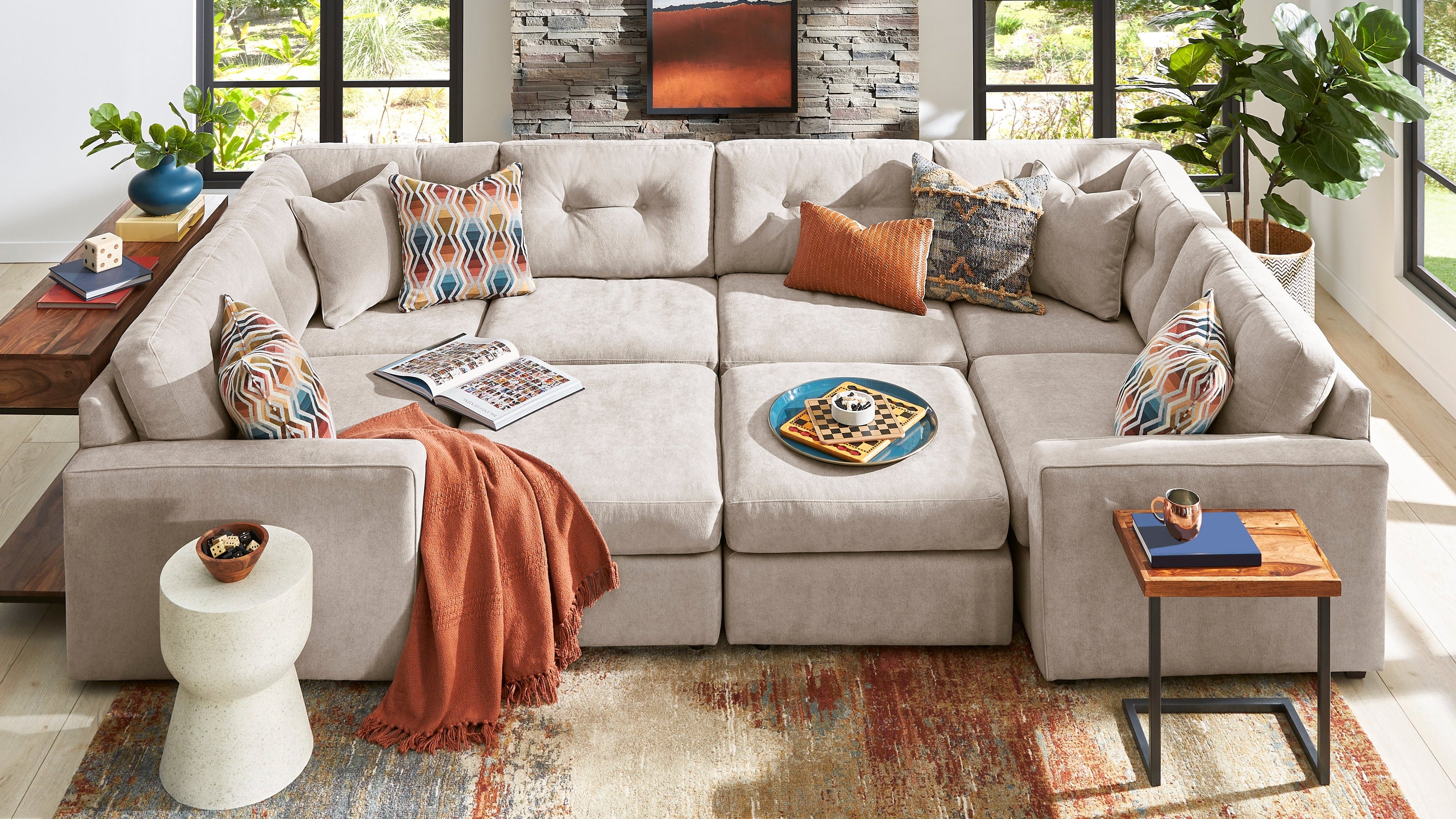 A light beige sectional sofa with multiple accent pillows and a rust-orange throw blanket, styled in a bright modern living room with large windows, a stone accent wall, and a patterned area rug.