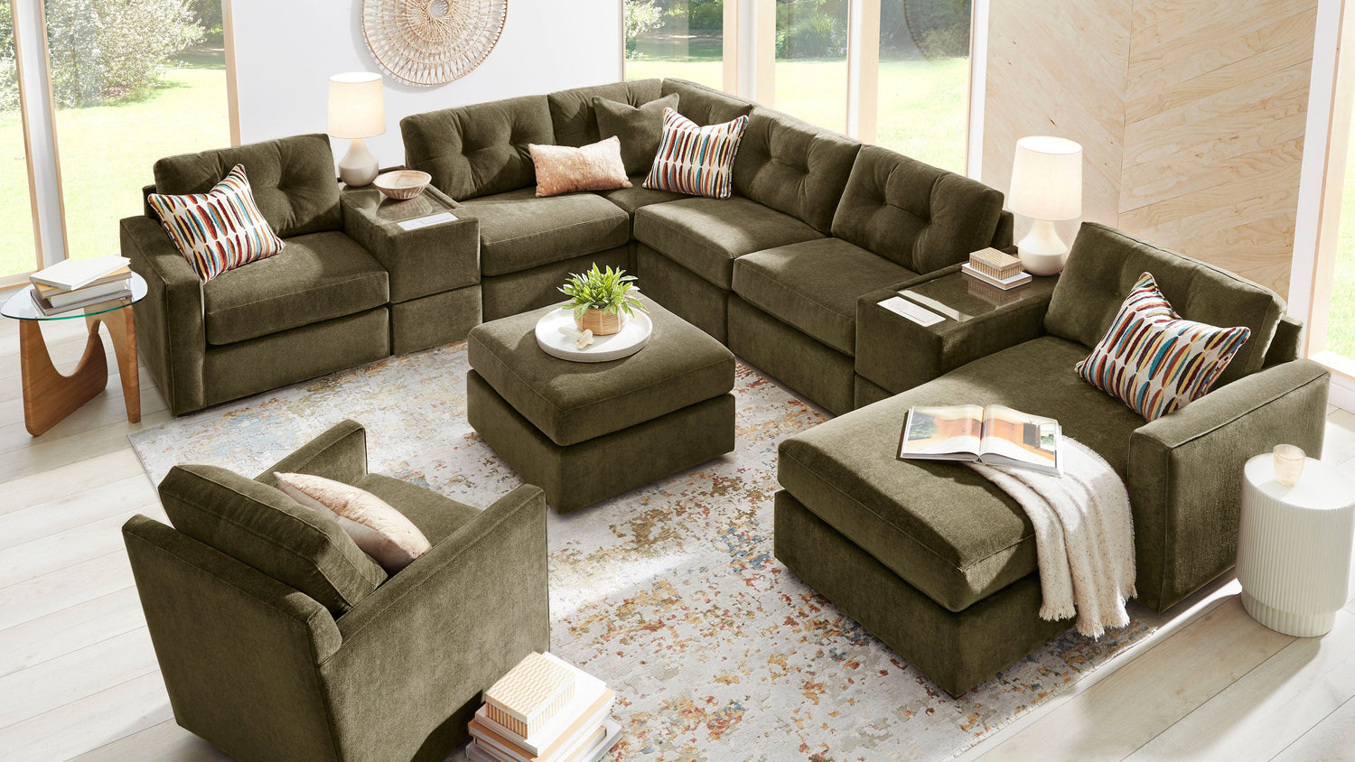An olive green sectional sofa set arranged in a bright, modern living room with large windows, light wood floors, and a patterned area rug. The set includes an L-shaped sectional, a matching armchair, a central ottoman, and striped accent pillows.