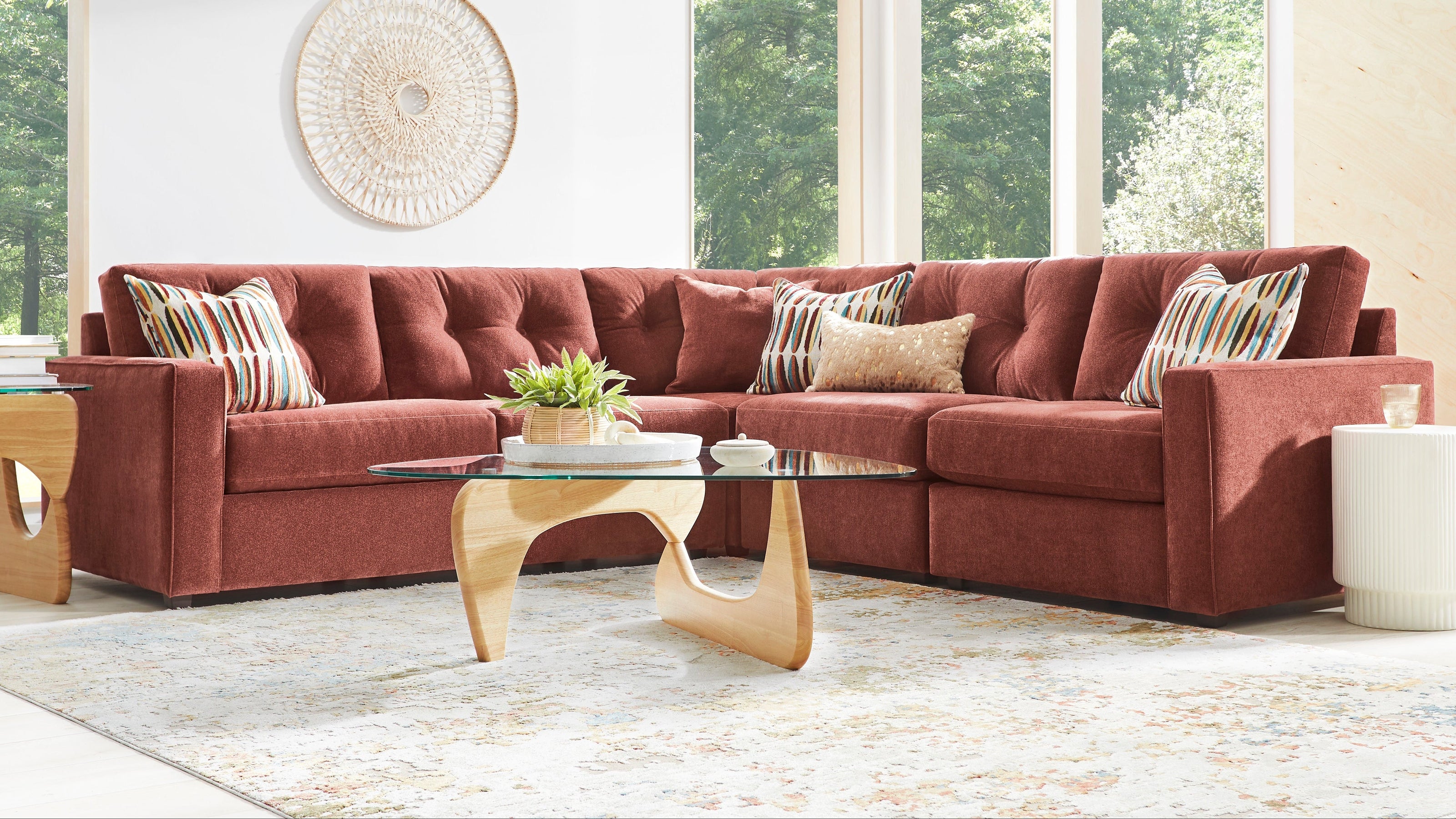 A modern sectional sofa upholstered in rust red fabric, styled in a bright living room with a patterned area rug, wooden coffee table, and large windows showing green trees outside.