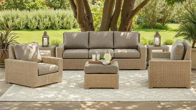 Outdoor patio furniture set with a sofa, chairs, and ottoman on a stone patio with trees and greenery in the background.