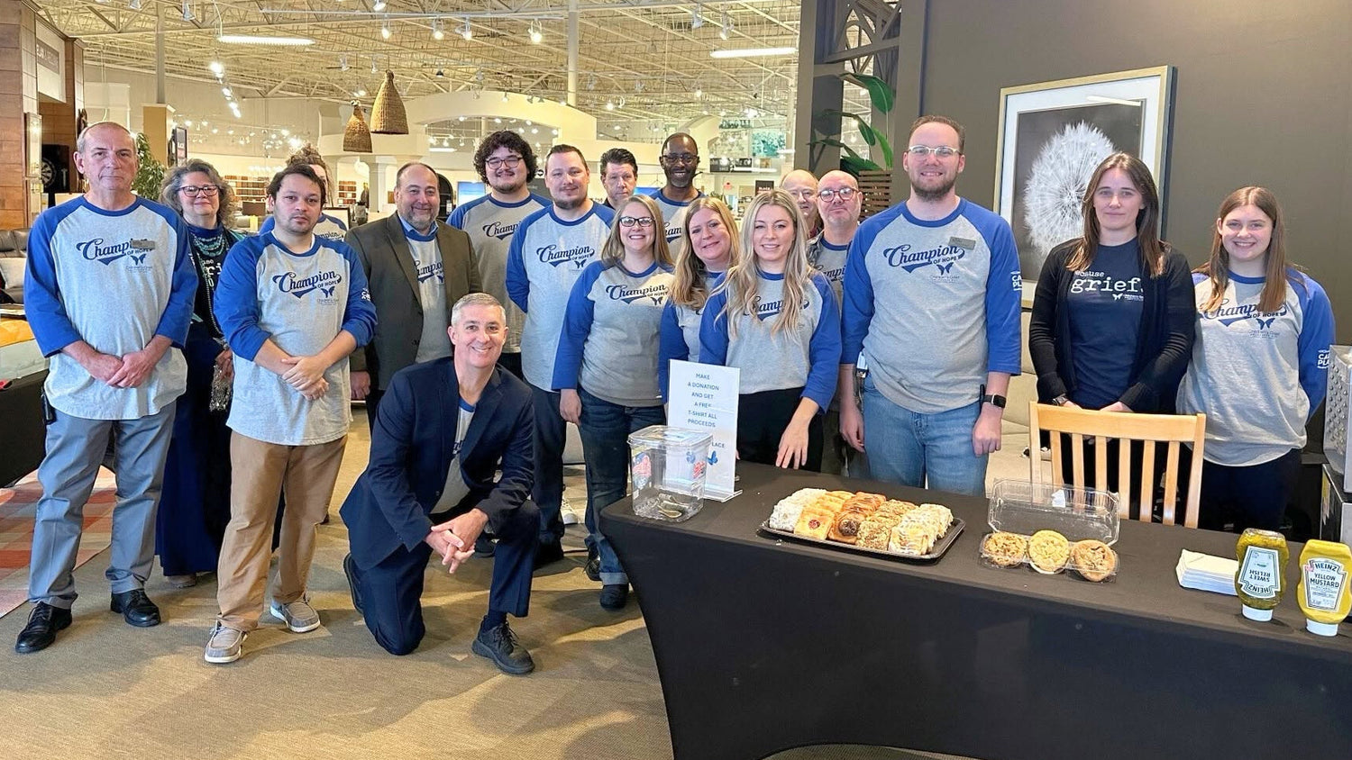 A group of people posing together in a retail showroom, wearing matching blue and gray raglan t-shirts. In the foreground, a black draped table holds food items and small bottles, with a donation sign standing beside it.