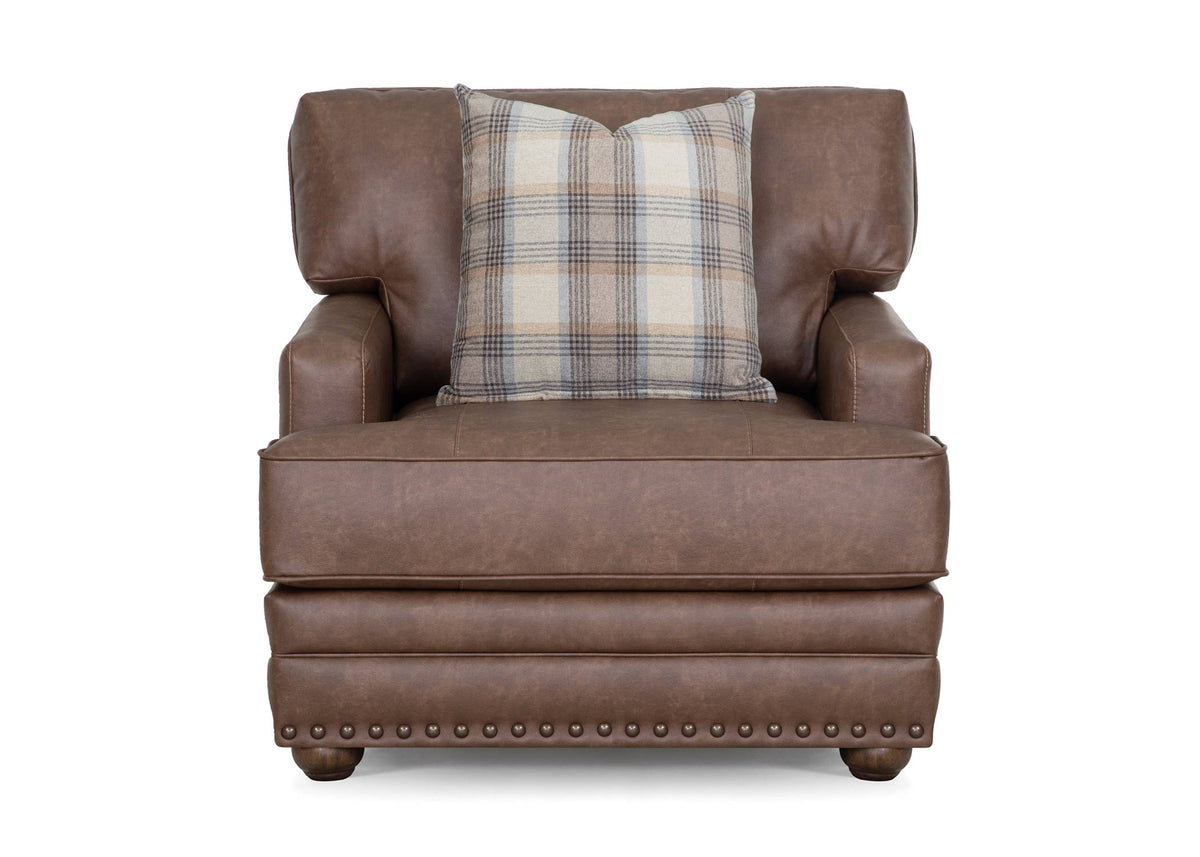 Brown upholstered accent chair with plaid pillow and nailhead trim along front skirt