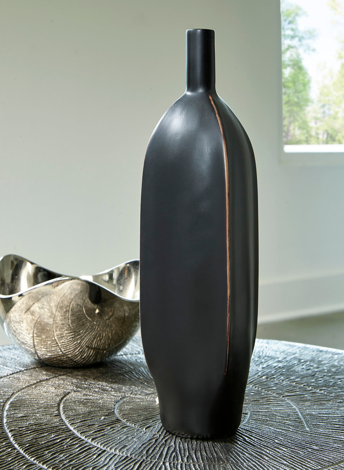 Black ceramic vase with tall neck on table surface, copper-toned vertical seam.