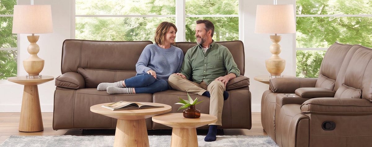 A man and woman relax on a brown leather sofa in a bright, modern living room, with a matching brown recliner chair to the right, wooden coffee tables, and table lamps on either side.