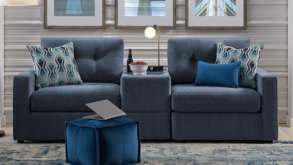 A modern living room setup featuring a dark blue tufted three-seat sofa, a matching armchair, a blue ottoman, and a blue patterned area rug. The furniture is styled against a silver horizontal-striped wall with three framed art prints.