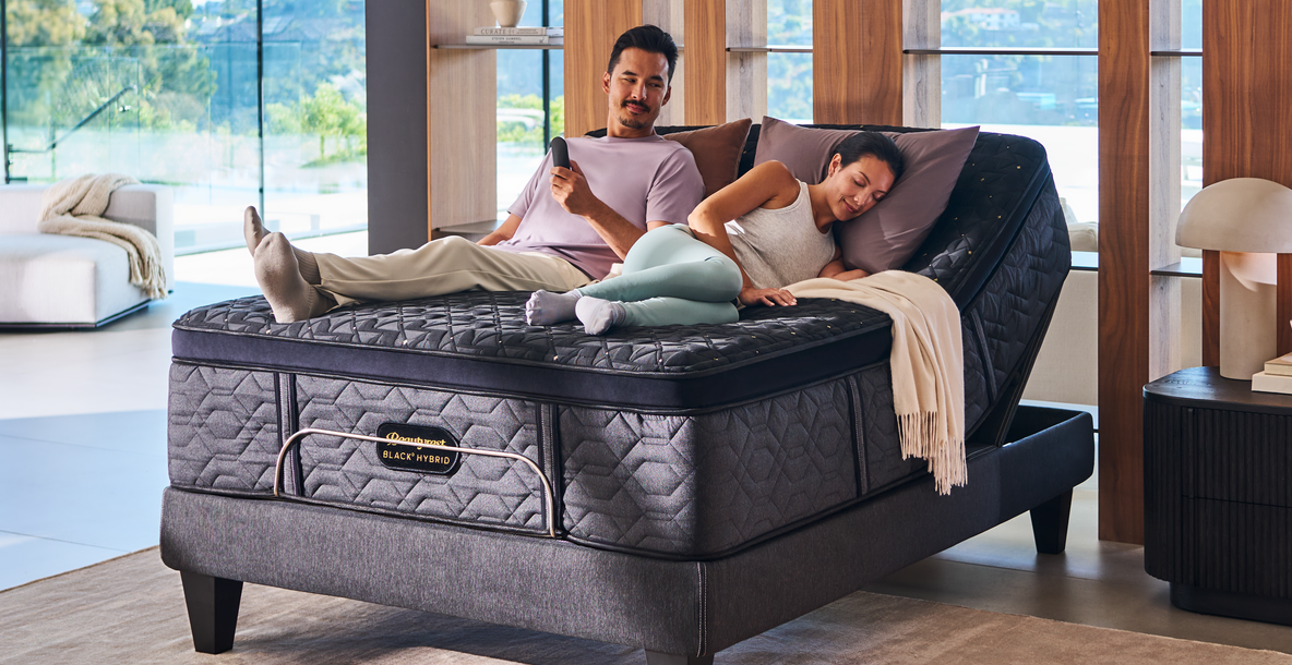 Man and woman lying on a gray adjustable bed in a modern room with large windows.