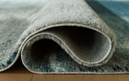 Area rug corner close-up showing rolled edge and blue-gray textured pile.